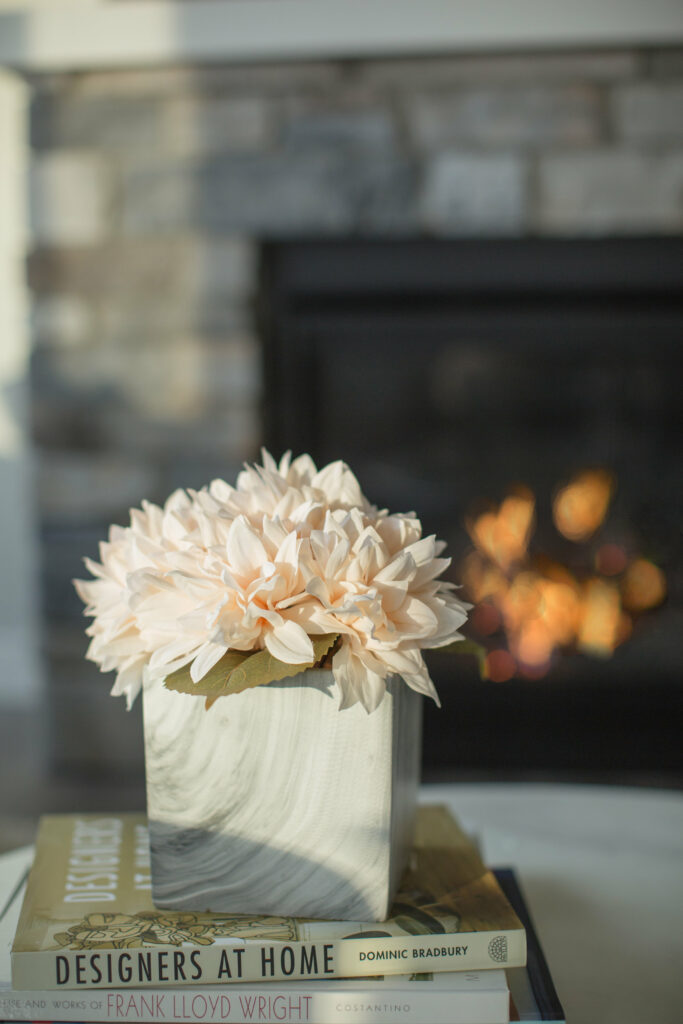 A staged living room with fireplace showing peach dahlias in a container ontop of books on a coffee table