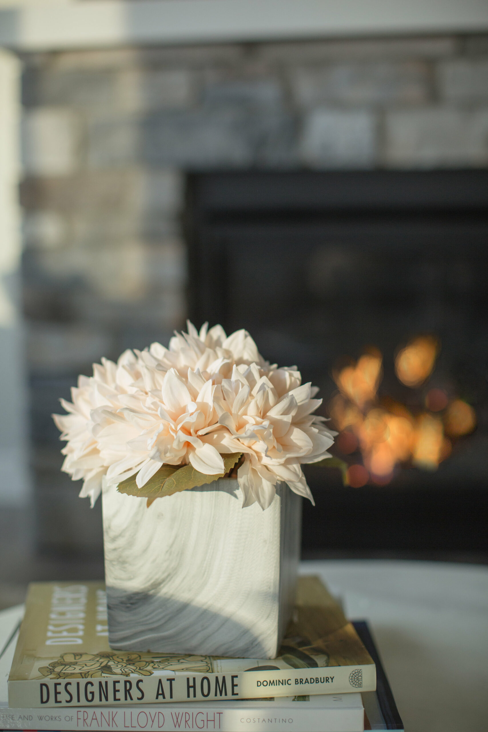 A staged living room with fireplace showing peach dahlias in a container ontop of books on a coffee table